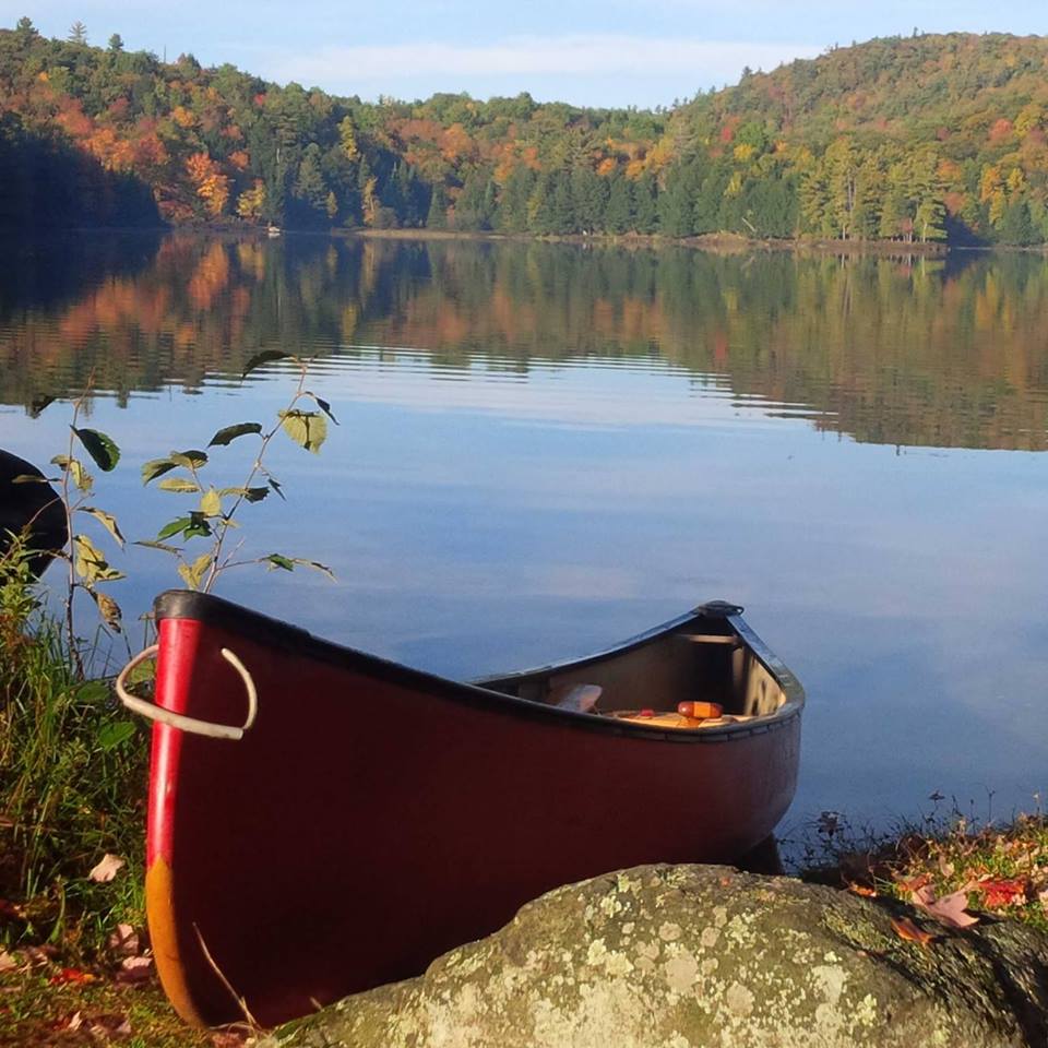 A scenic view of lake, forest drapped in fall colours and a red canoe on the shore.